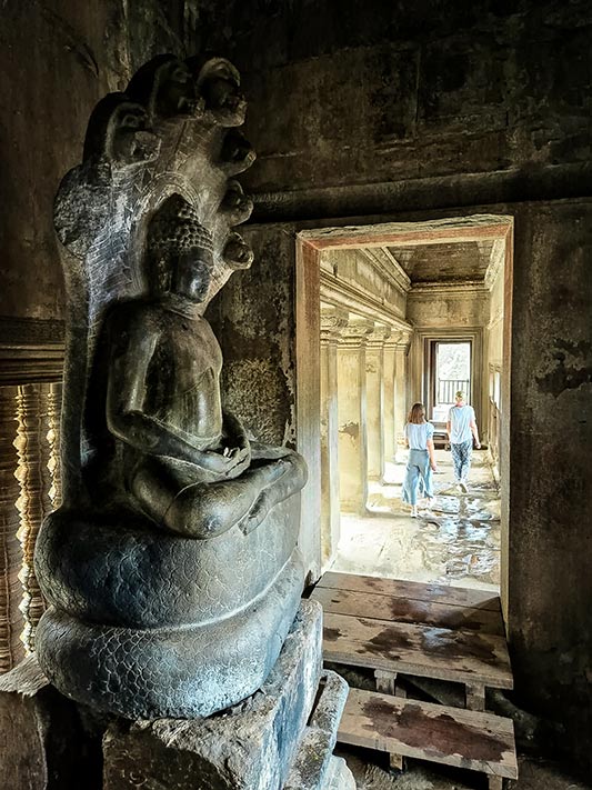 Vorn ein Buddha auf einer Naga sitzend in Angkor Wat. Hinten gehen meine Tochter und ich durch die Galerie.